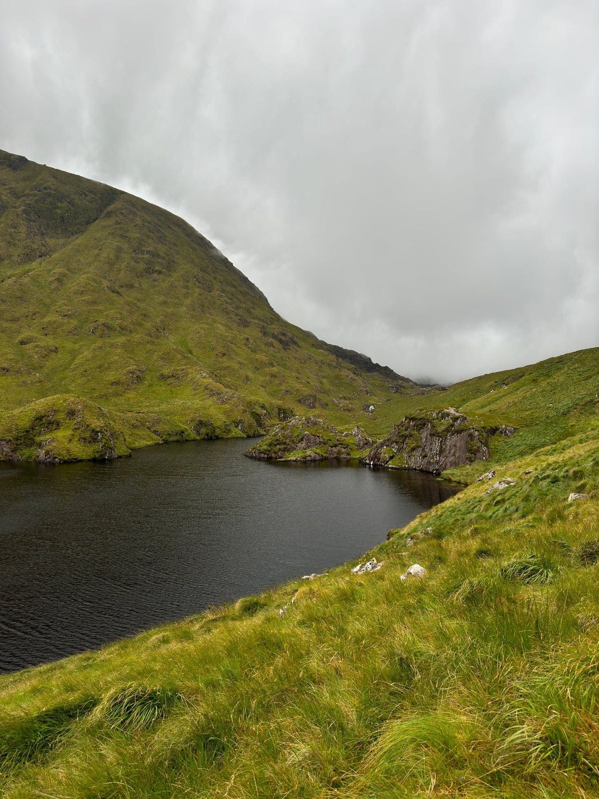 A dark mountain lake enclosed by steep green slopes and low cloud