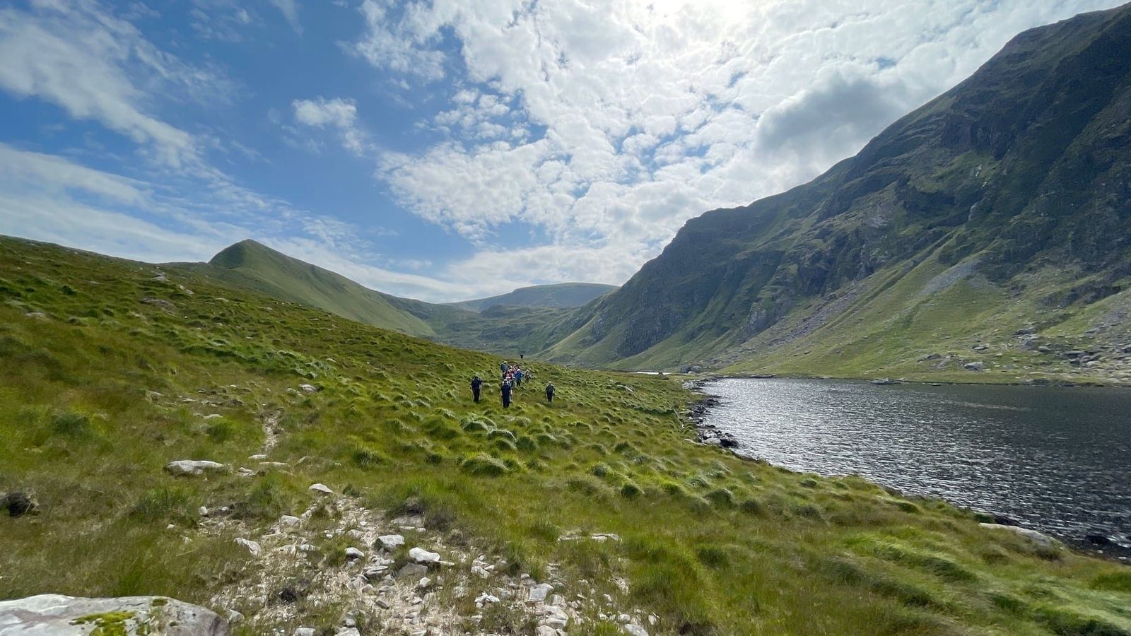 Walkers crossing rough ground beside a mountain lake in the Com an Áir corrie