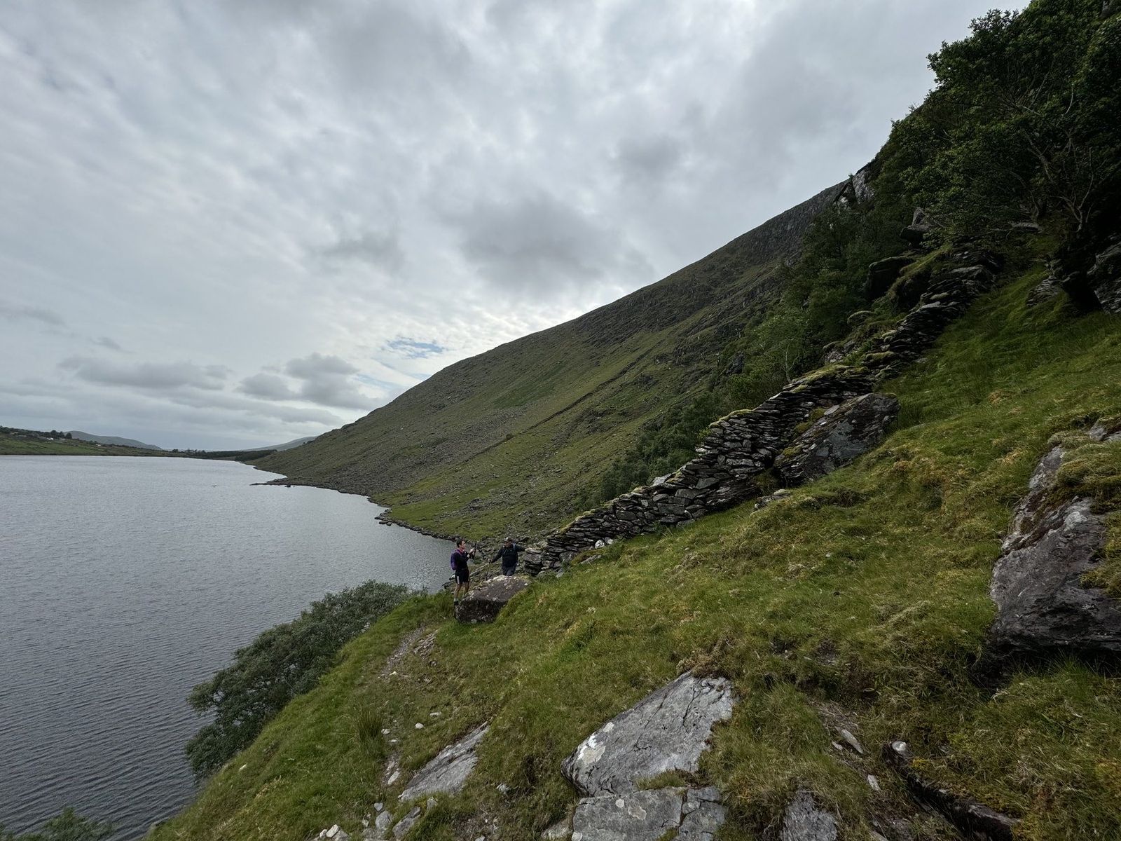 Walkers traversing steep green ground beside a mountain lake