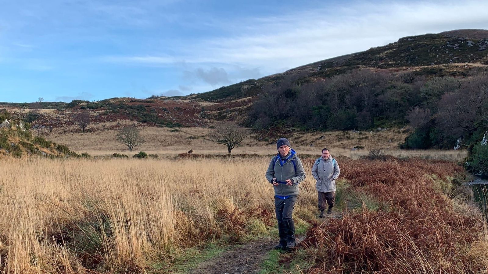 Two walkers crossing open ground below wooded hills