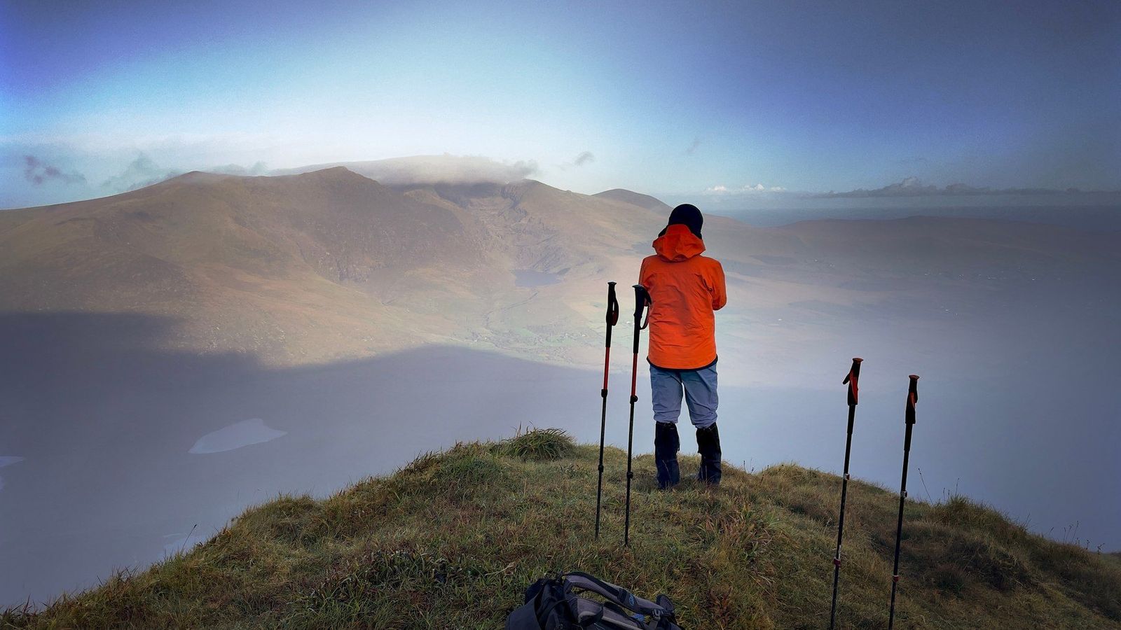 A hillwalker in an orange jacket standing on a grassy crest above cloud