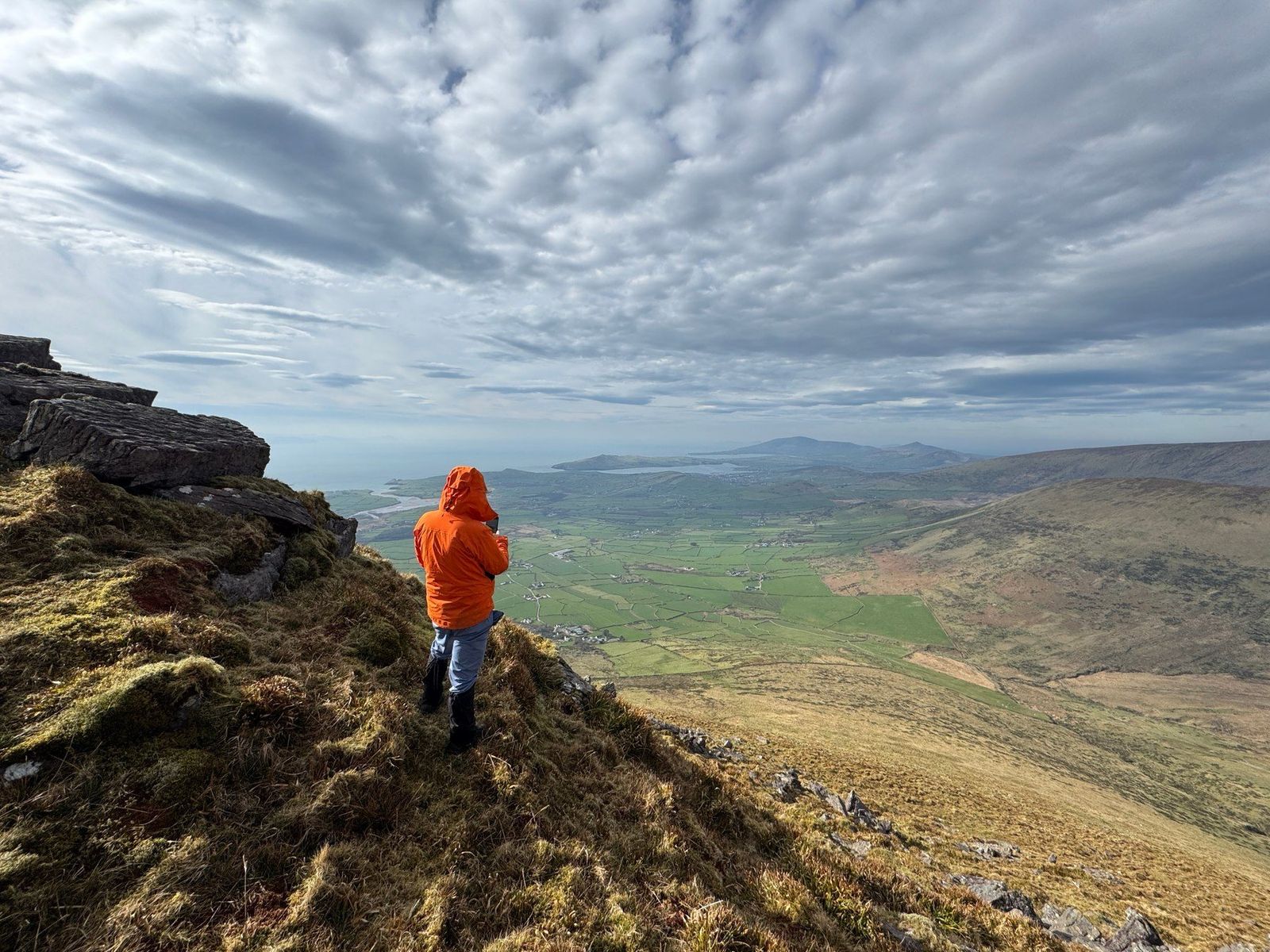 A walker descending rough ground towards green fields far below