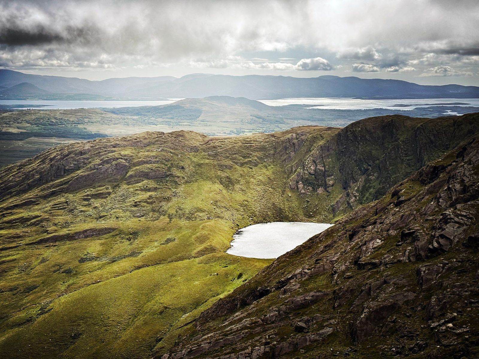A wide mountain panorama over a bright corrie lough towards the Kerry coast