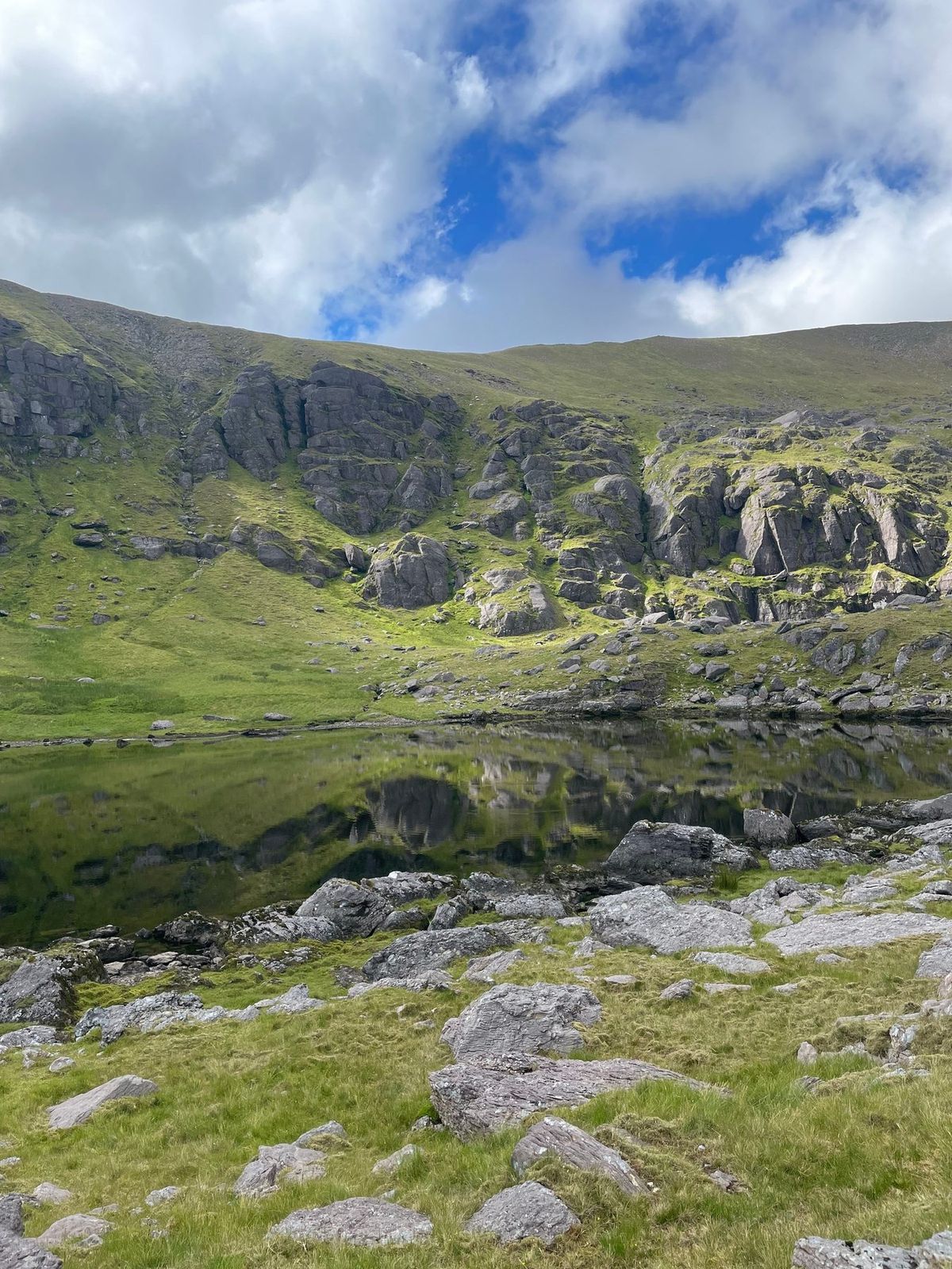 A dark corrie lough reflecting broken cliffs and bright mountain grass