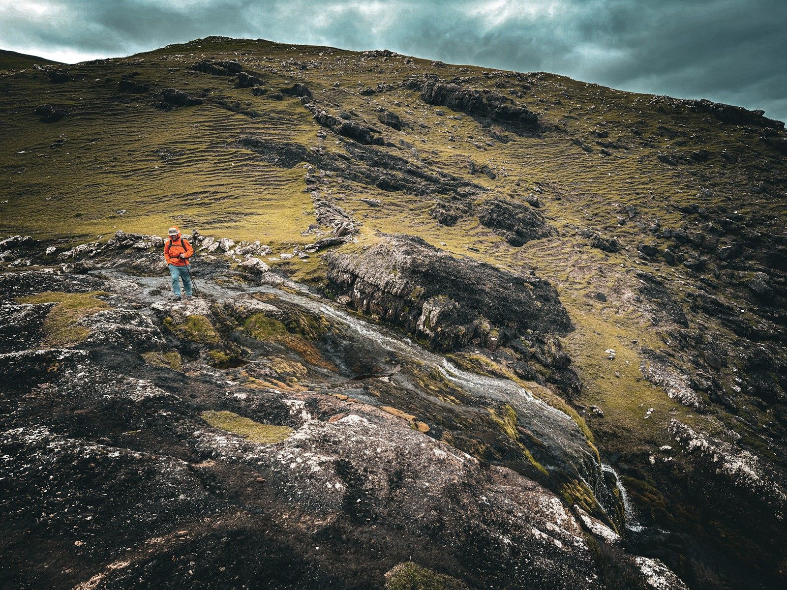 A hillwalker in an orange jacket crossing wet rock beside a coastal cascade above the sea cliffs