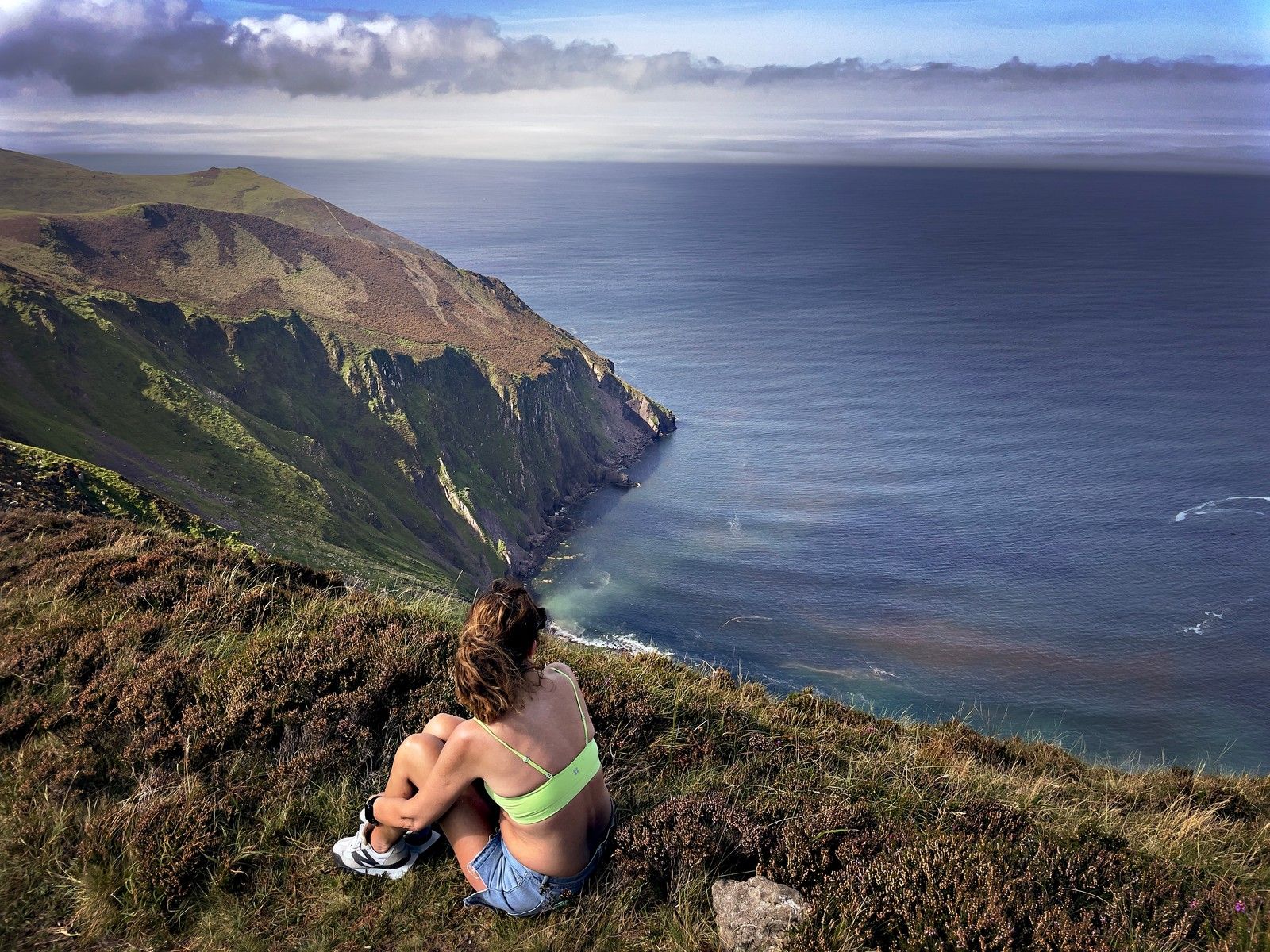 A walker resting on heather above the high cliffs and calm Atlantic water